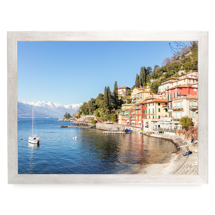 A picturesque coastal town with colorful buildings, a sandy beach, and a body of water with a sailboat. The town is situated on a hillside overlooking the water, with snow-capped mountains visible in the distance.