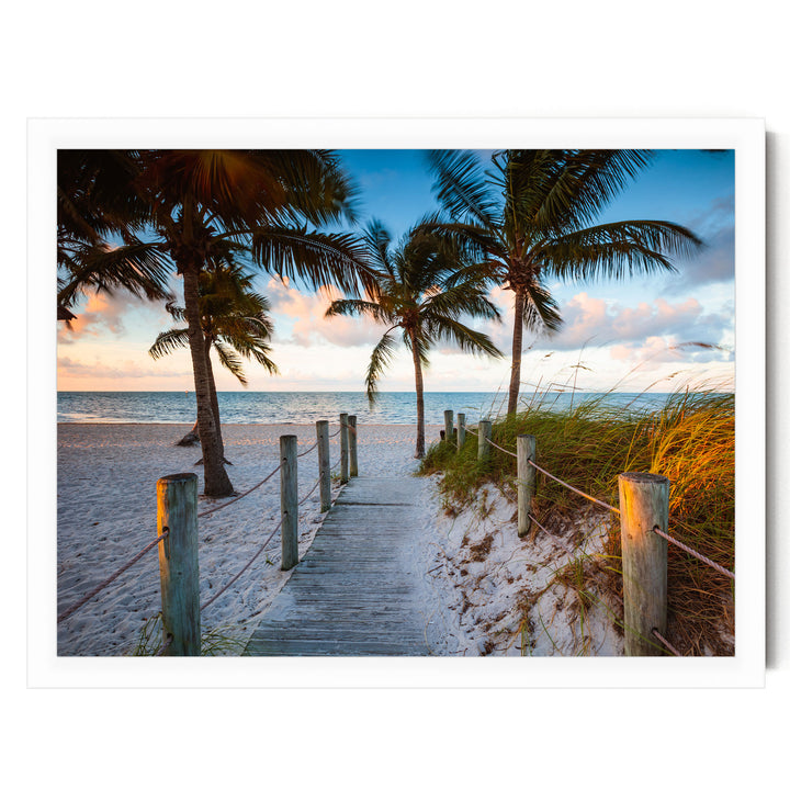 A wooden walkway leads to a sandy beach, lined with palm trees and a fence, with a clear blue sky and colorful sunset in the background.