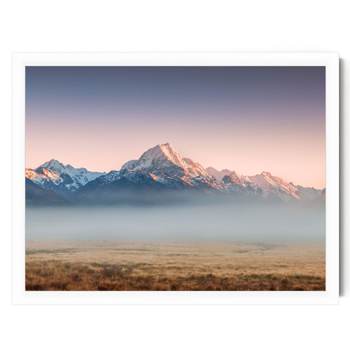 A breathtaking landscape featuring snow-capped mountains in the distance, with a hazy, golden-hued sky above. The mountains are partially obscured by a misty haze, creating a serene and atmospheric scene.