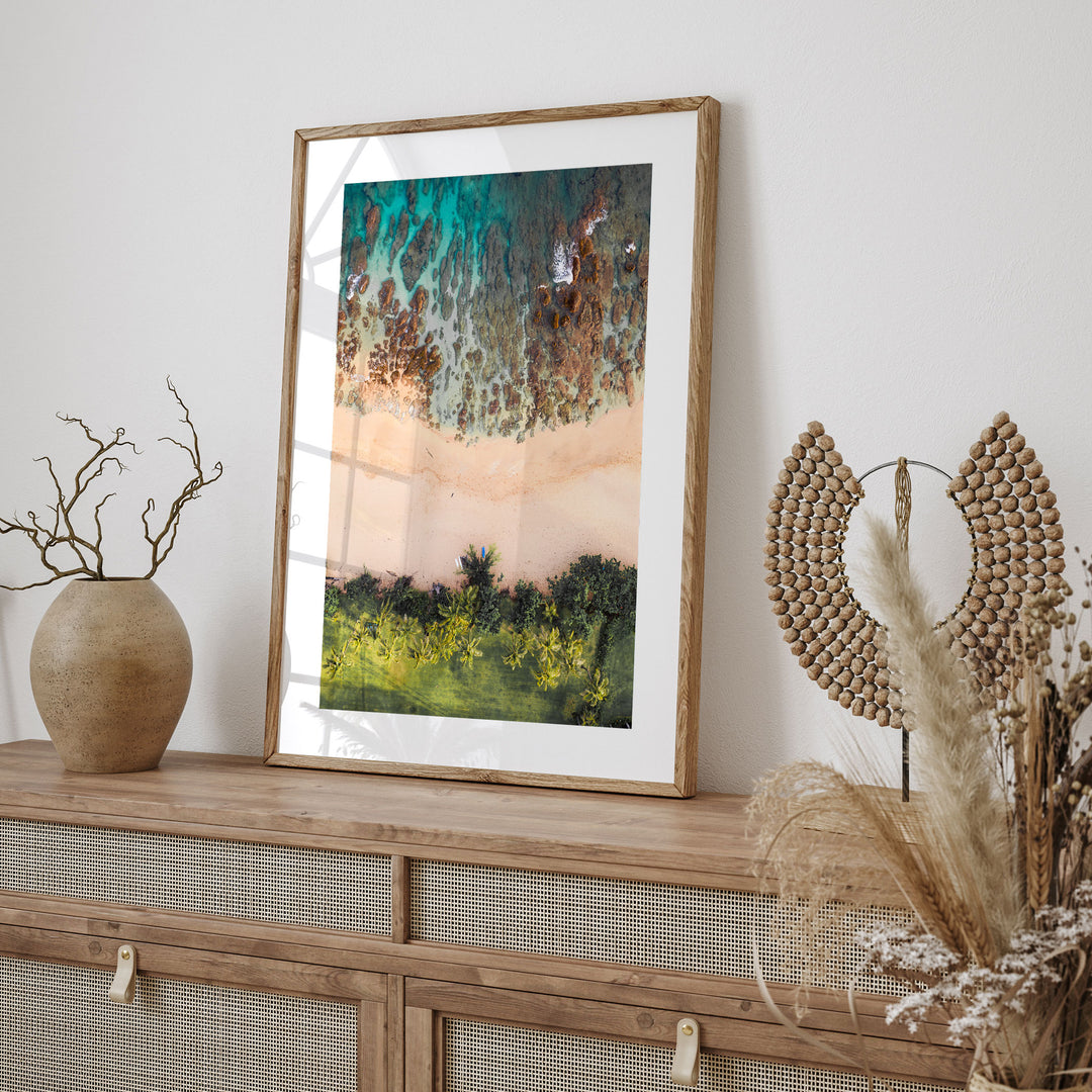 a framed painting of a beach scene with a mix of green and brown colors, hanging on a wall above a wooden cabinet or shelf.