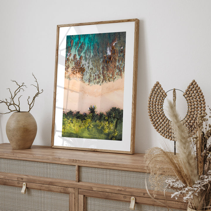 a framed painting of a beach scene with a mix of green and brown colors, hanging on a wall above a wooden cabinet or shelf.