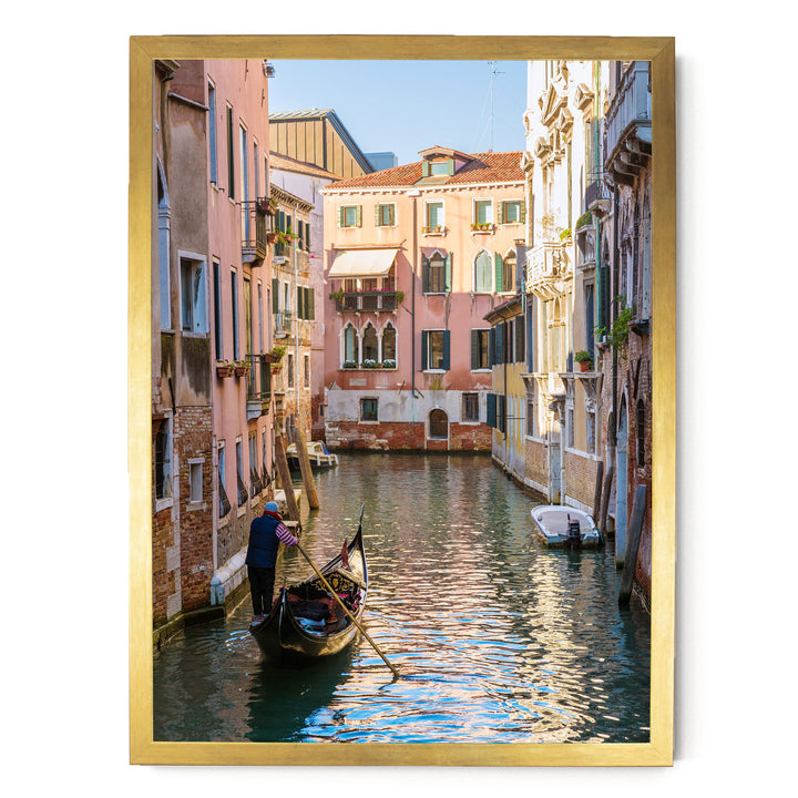 A gondolier in a traditional Venetian gondola navigates a picturesque canal in Venice, surrounded by colorful buildings and reflecting the clear blue sky.