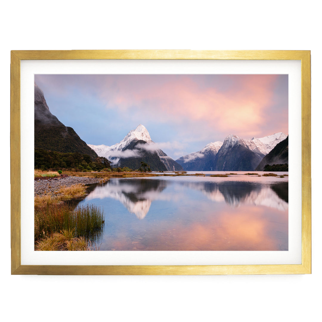 a serene landscape with a calm lake reflecting the surrounding mountains and sky. The mountains are covered in snow, and the sky is a beautiful gradient of pink and orange, creating a stunning backdrop for the scene. The lake is nestled amidst lush vegetation, adding to the overall tranquility of the image.