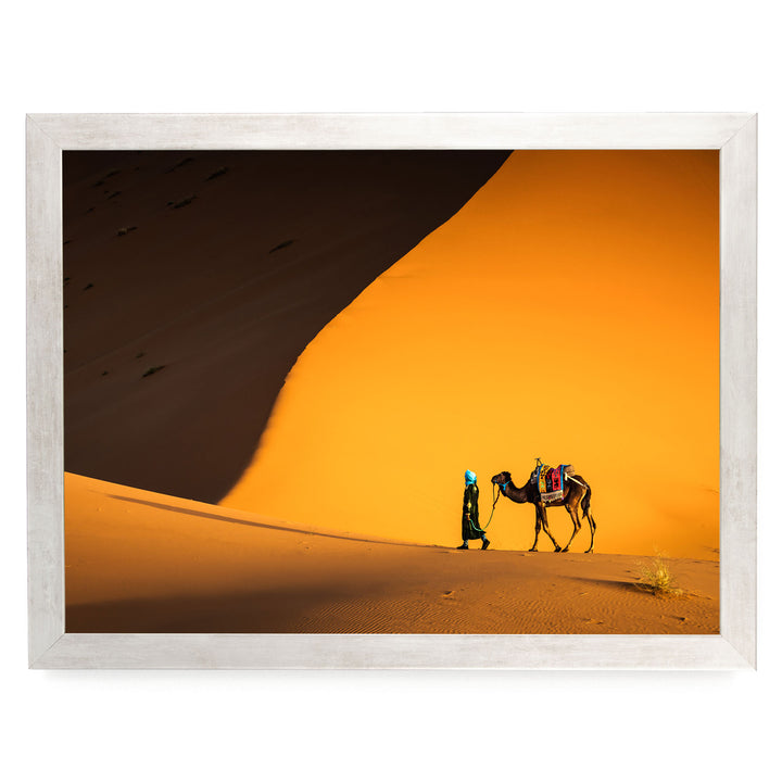 A person in a green outfit is walking a camel across a sandy desert landscape with a large sand dune in the background.