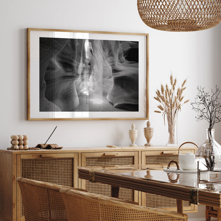 a dining room with a large framed black and white photograph hanging on the wall above a wooden cabinet. The cabinet has drawers and a glass top. The dining table is set with a white teapot and a vase of flowers. The room has a rustic, natural aesthetic with woven furniture and a woven light fixture hanging from the ceiling.
