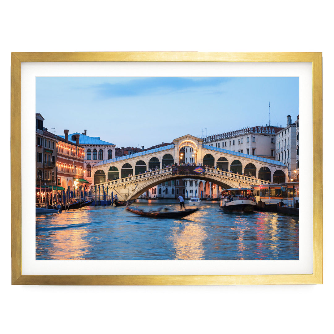 a picturesque scene of the Rialto Bridge in Venice, Italy, with its distinctive white stone arch spanning the Grand Canal. The bridge is illuminated by warm lights, creating a romantic atmosphere. The canal is filled with boats, adding to the lively ambiance of the scene. The image is framed in a gold frame, enhancing its visual appeal.
