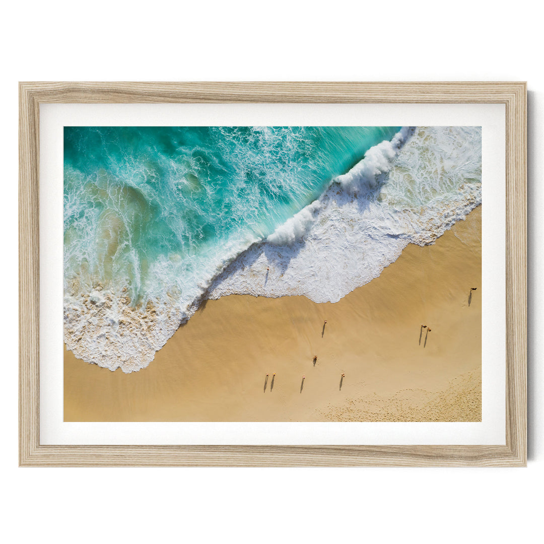 The image is a framed photograph of a beach scene with people walking along the shoreline, where the waves are crashing onto the sand.