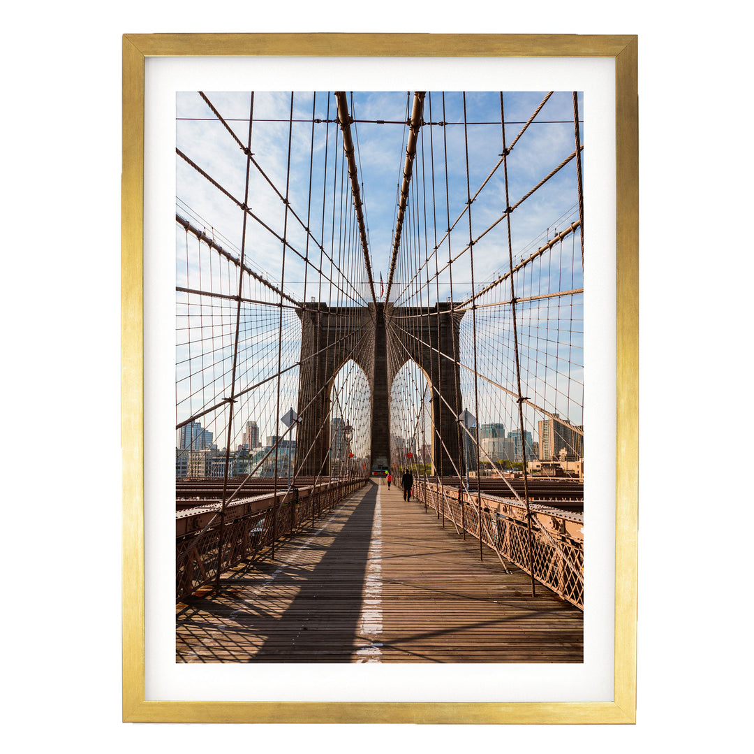 the iconic Brooklyn Bridge, with its distinctive arches and suspension cables, spanning across the frame. The bridge is surrounded by a cityscape, with tall buildings visible in the background. The photograph is framed in a gold frame, adding a touch of elegance to the scene.