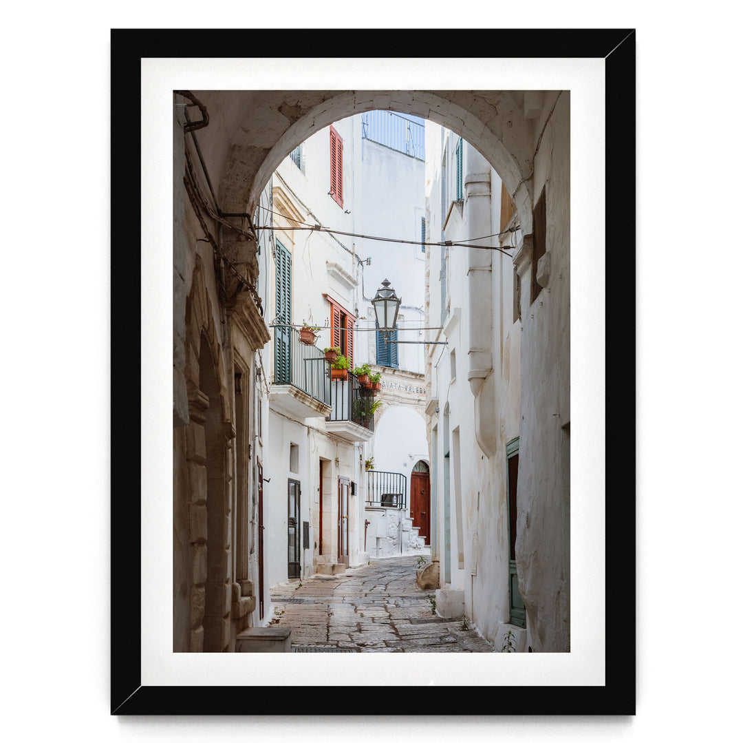 A narrow cobblestone street lined with white buildings and colorful shutters, leading to a balcony with potted plants.