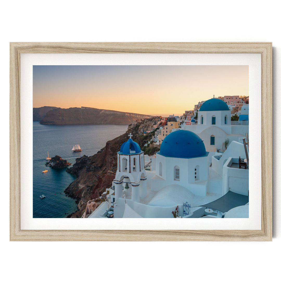 a picturesque coastal town with white buildings and blue domes, overlooking the ocean. The town is situated on a cliff, with a clear blue sky and a warm sunset in the background.