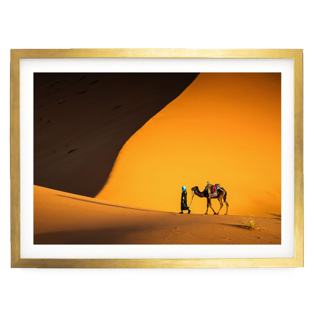 A person is walking with a camel in a desert setting, with a large sand dune in the background.