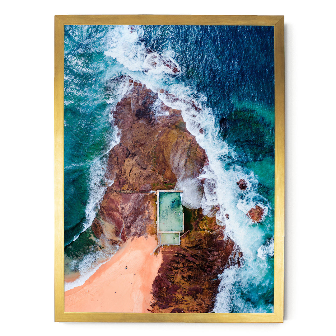 The image is an aerial view of a beach with a swimming pool, surrounded by rocky cliffs and crashing waves.