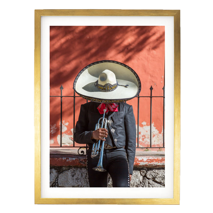 A person wearing a sombrero and a black jacket is playing a trumpet in front of a red wall.