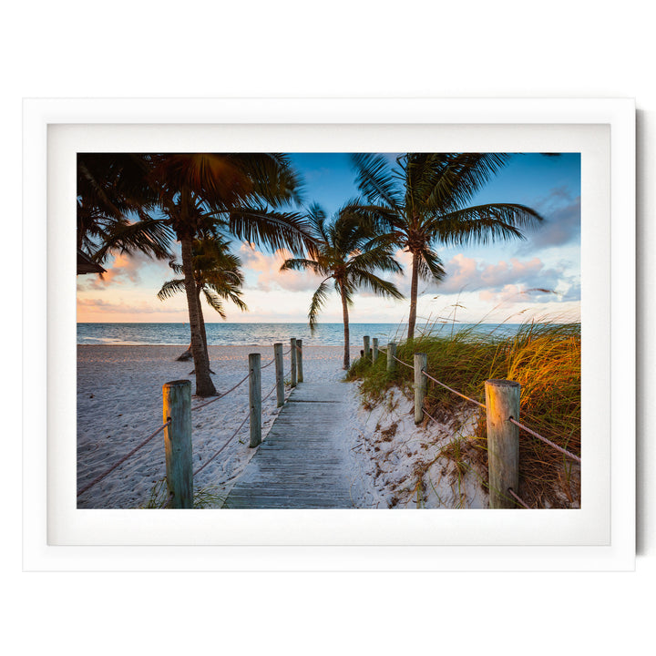 A wooden walkway leads to a sandy beach, with palm trees and a colorful sunset sky in the background.