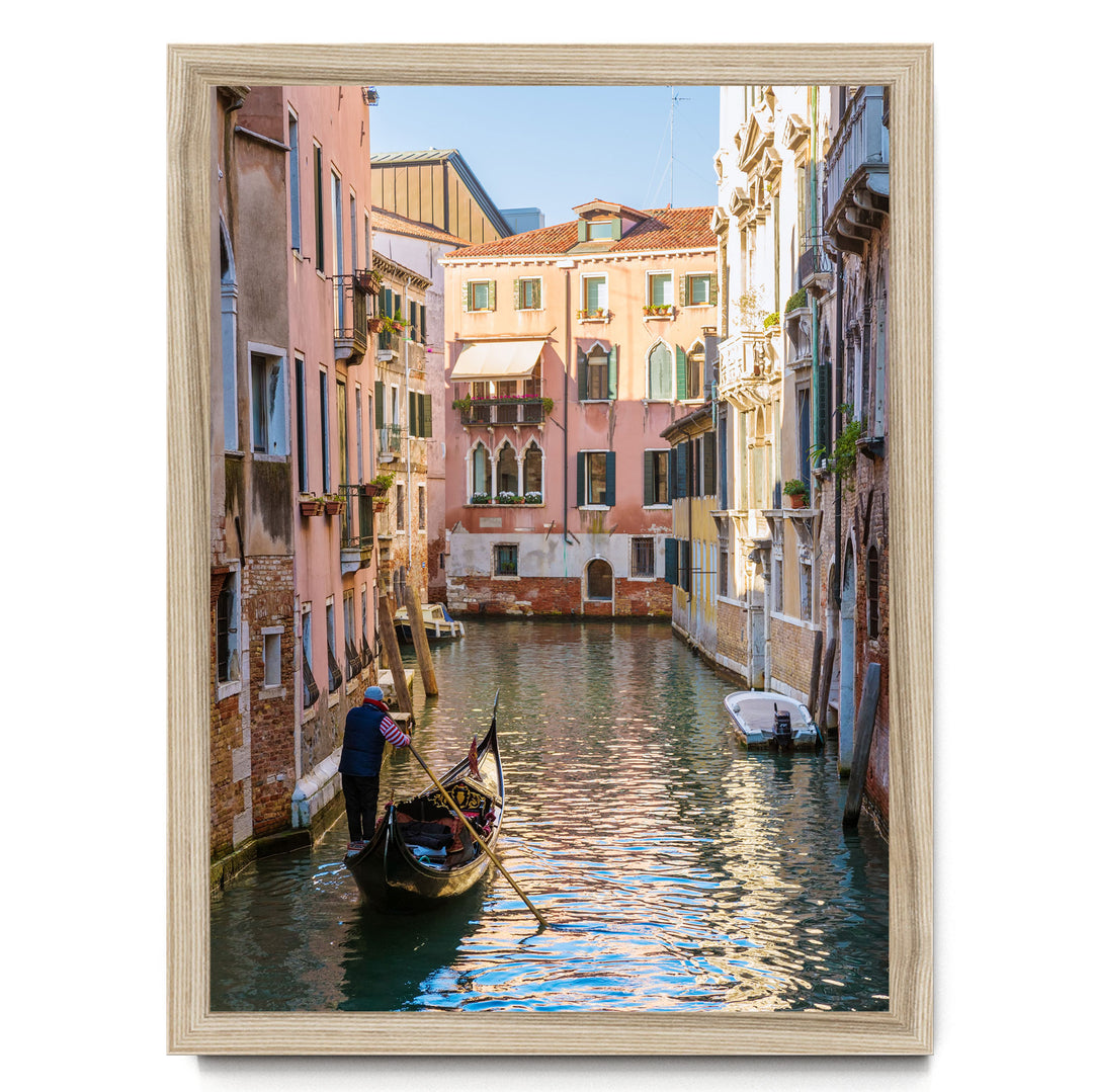 A gondolier in a traditional Venetian gondola navigates a picturesque canal in Venice, surrounded by colorful buildings and reflecting the clear blue sky.