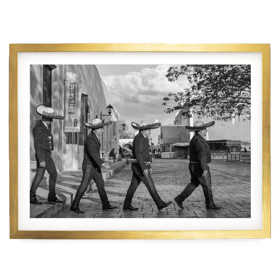 A black and white photograph of four men wearing sombreros walking in a line on a brick street, with a building and trees in the background.