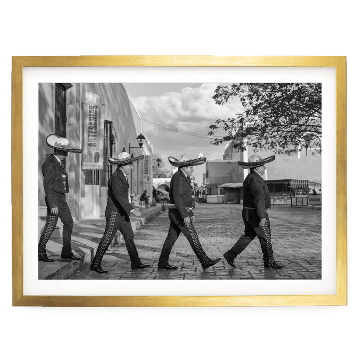 A black and white photograph of four men wearing sombreros walking in a line on a brick street, with a building and trees in the background.