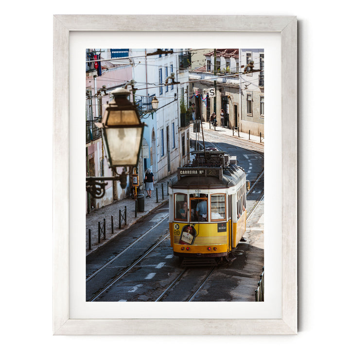 A yellow tram is traveling down a street in a European city, with buildings and pedestrians visible in the background.