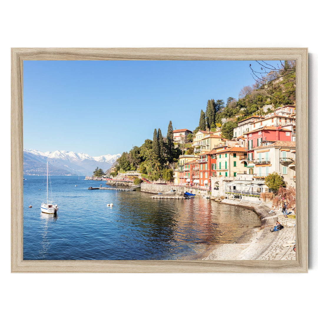 a picturesque coastal town with colorful buildings lining the shoreline, overlooking a calm lake. A sailboat is visible on the water, and the town is nestled among lush green hills.