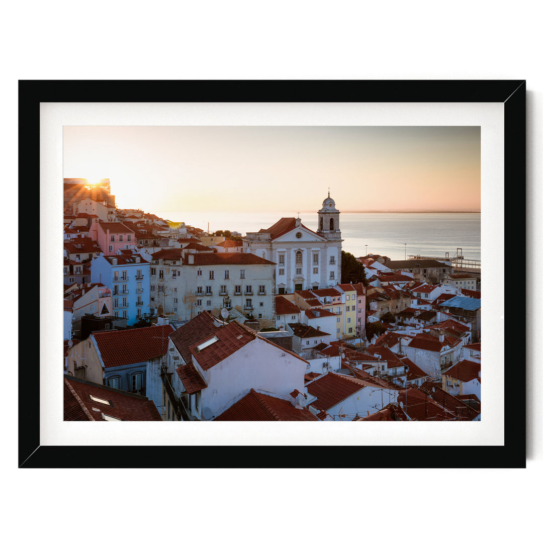 A picturesque view of a coastal town with colorful buildings, a church with a bell tower, and the sun setting over the ocean in the distance.