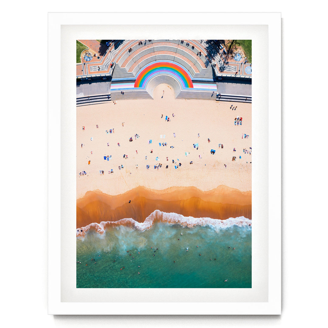 The image is an aerial view of a beach with a large, colorful arch structure in the background, and the ocean with waves crashing onto the shore.