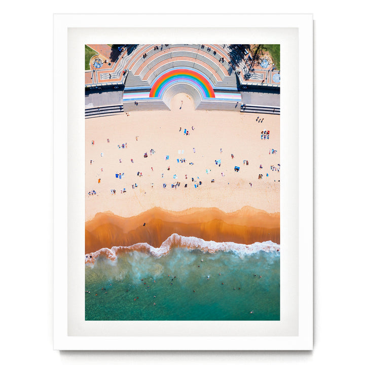 The image is an aerial view of a beach with a large, colorful arch structure in the background, and the ocean with waves crashing onto the shore.