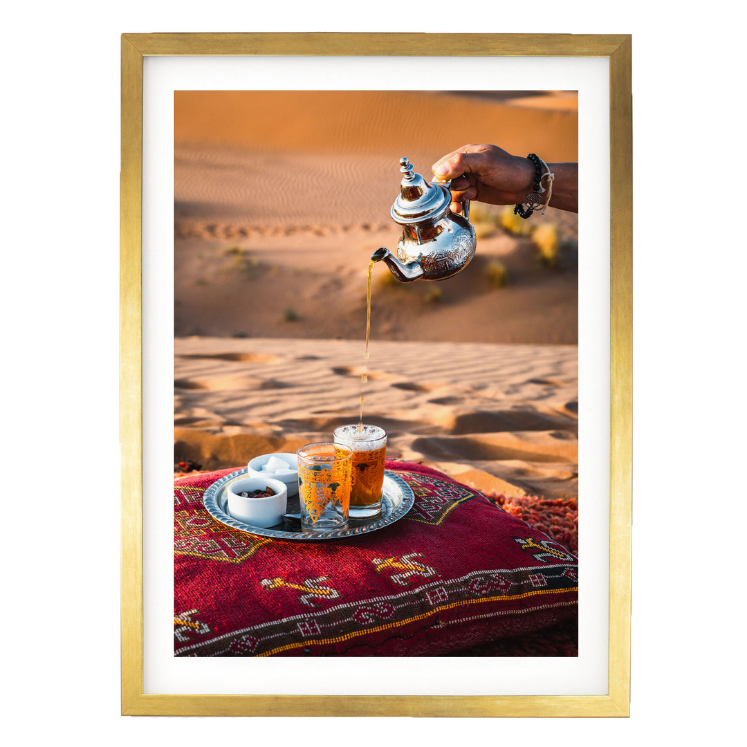 A person is pouring tea from a silver teapot into a glass, with a tray of tea cups and saucers on a red and gold patterned cushion, set against a desert landscape.