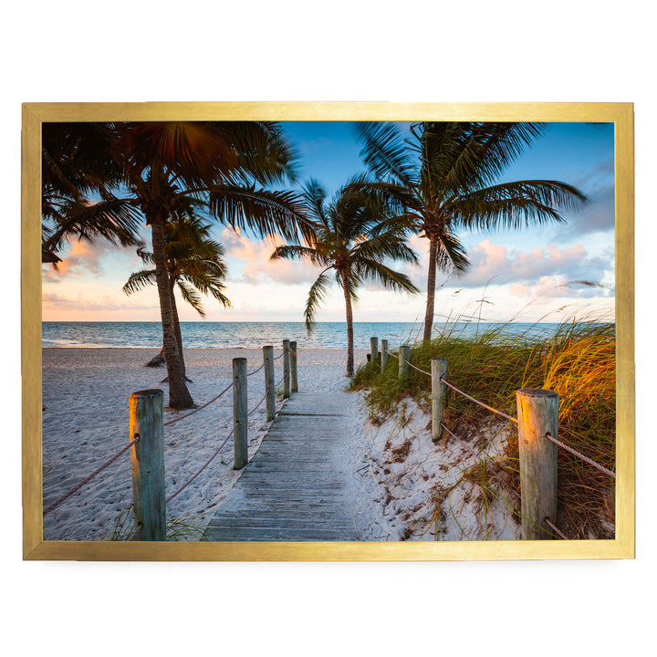 A wooden walkway leads to a sandy beach, with palm trees and a blue sky in the background.