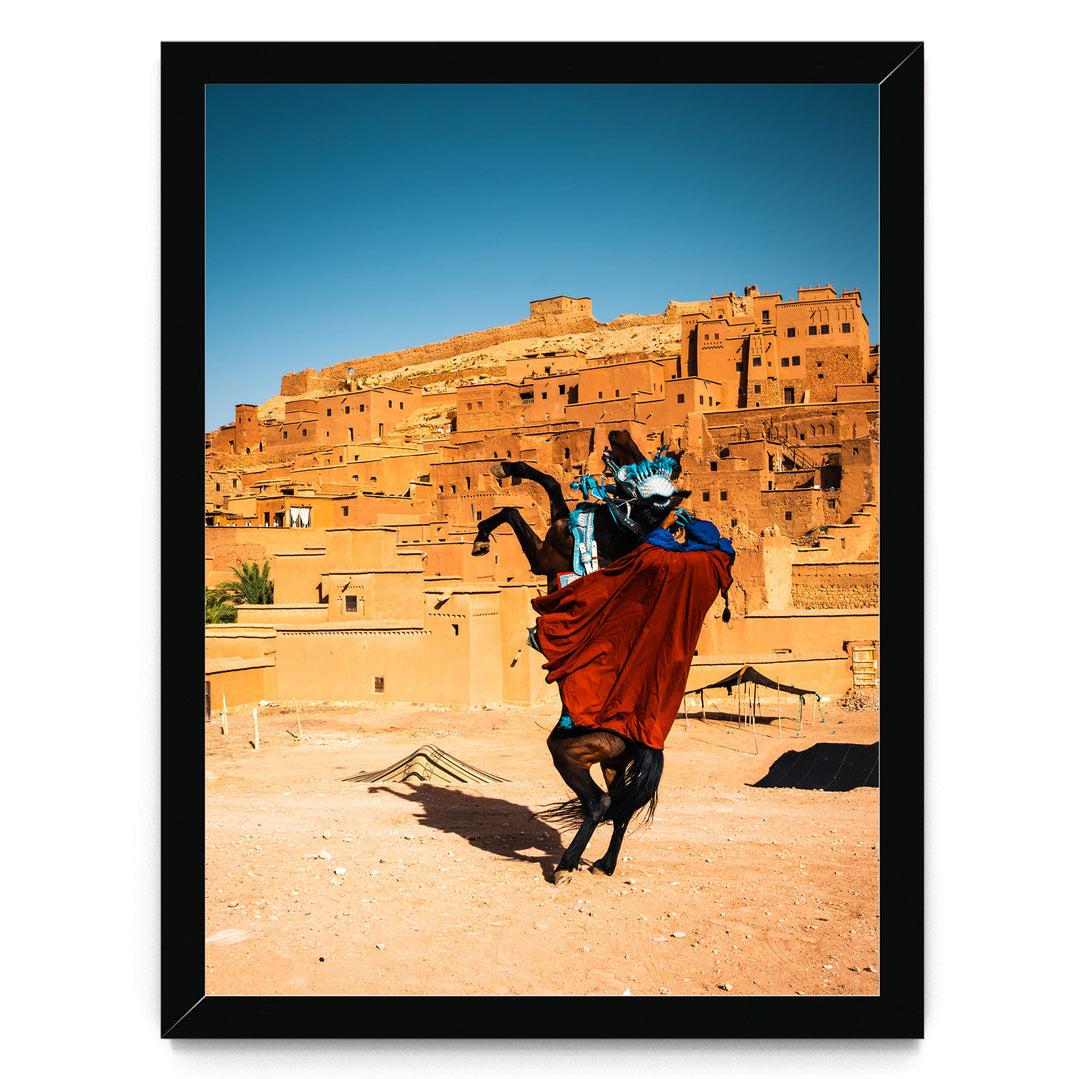A person in a red cape rides a horse in front of a desert-like landscape with orange-colored buildings in the background.