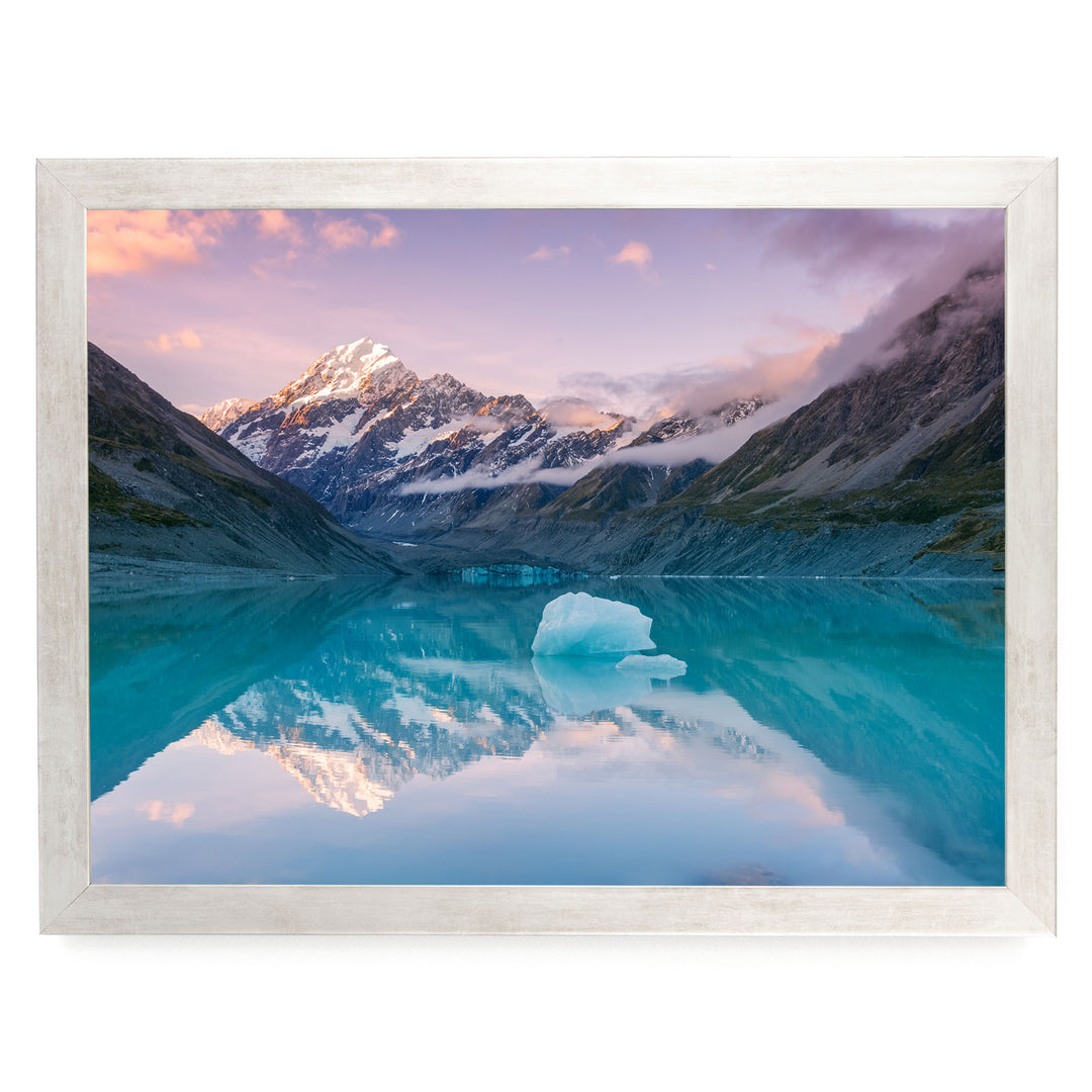 A breathtaking landscape featuring a serene lake with a large iceberg floating on the water, surrounded by majestic snow-capped mountains under a vibrant sky.