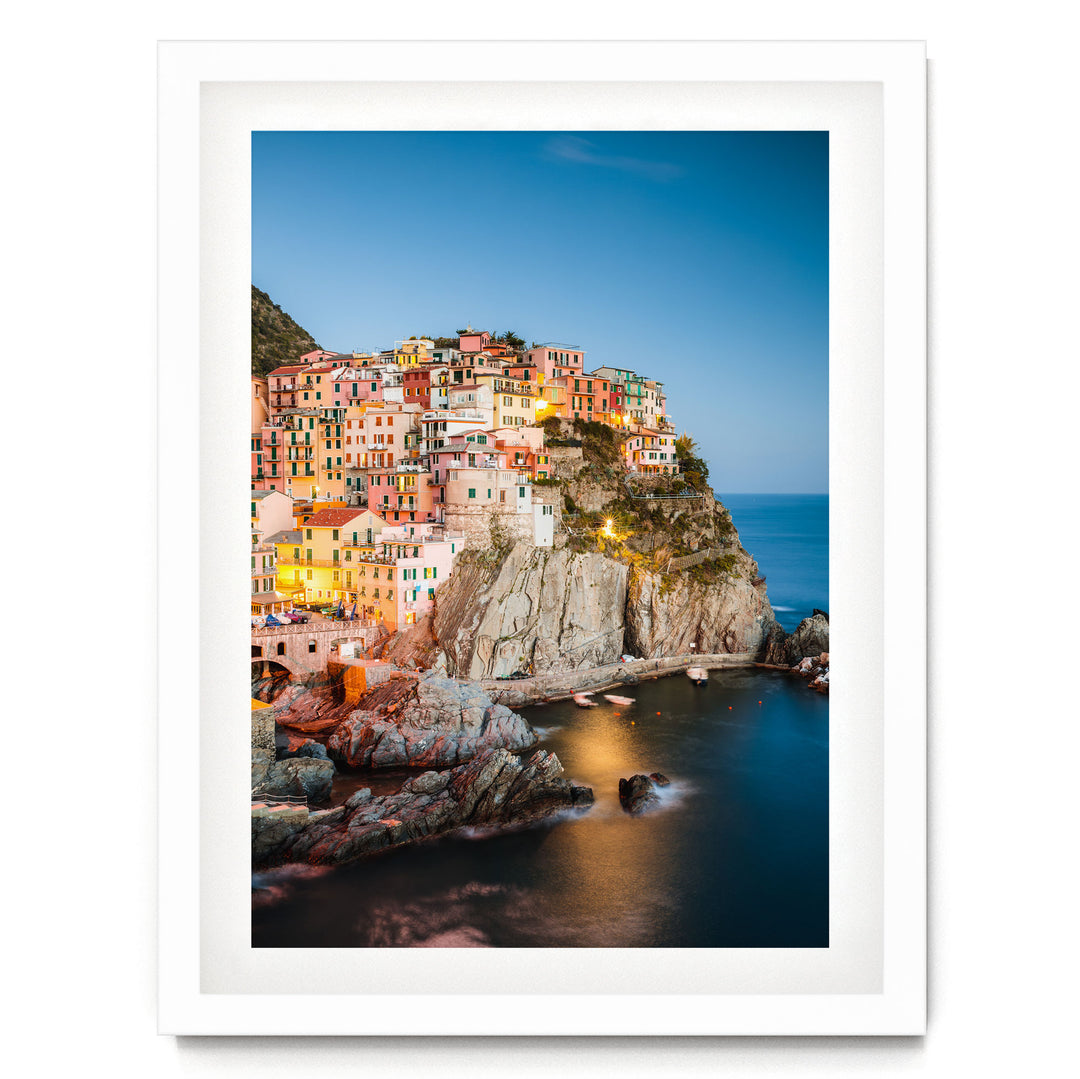 A picturesque coastal town nestled on a cliff, with its buildings illuminated by warm lighting against a clear blue sky.