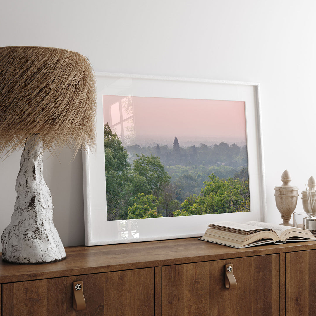 a framed picture of a forest landscape with a pink and orange sky in the background, placed on a wooden dresser next to a lamp.