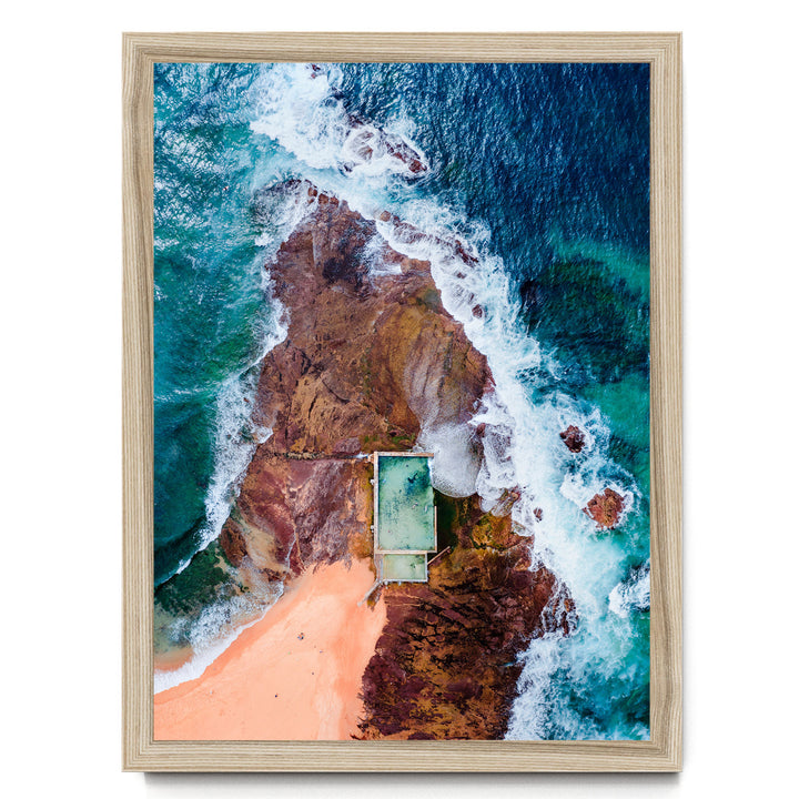 The image is an aerial view of a beach with a swimming pool, surrounded by rocky cliffs and crashing waves.