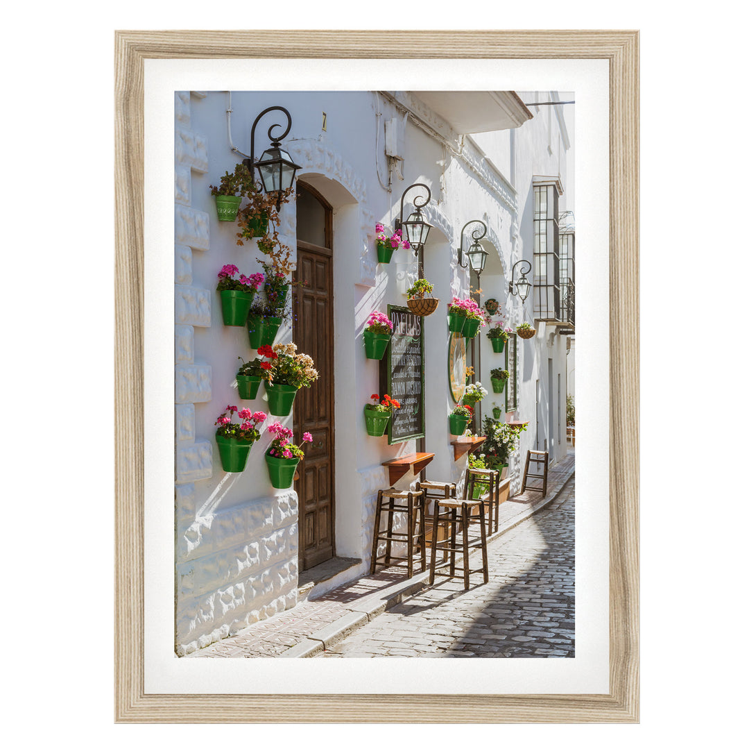 A charming street scene with a white building adorned with hanging flower pots and a wooden door. The building features a balcony with a railing and a small table and chairs. The cobblestone street and the sunlight create a warm and inviting atmosphere.
