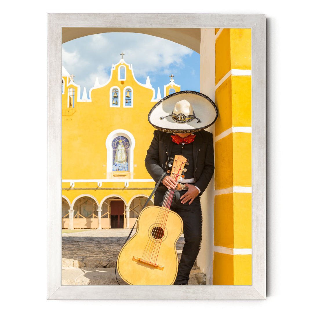 A person in a sombrero and traditional Mexican attire is playing a guitar in front of a yellow building with arched windows and a cross on top.