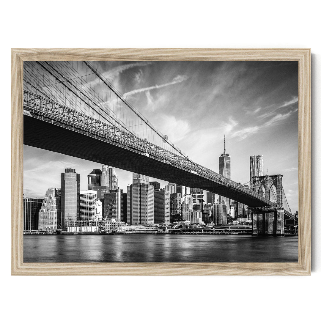 A black and white photograph of the Brooklyn Bridge spanning the East River, with the New York City skyline visible in the background.
