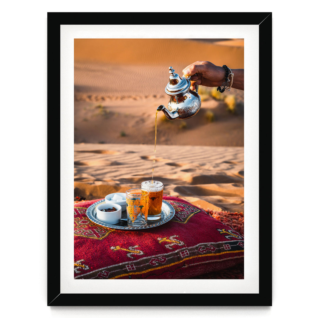 A hand pours tea from a silver teapot into a glass, with a tray of tea and snacks on a red and gold patterned cushion, set against a desert landscape.
