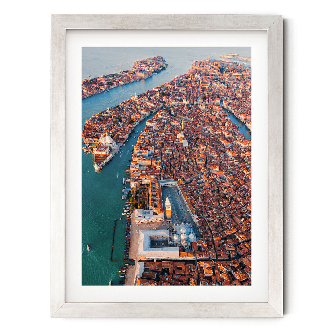 The image is an aerial view of a densely populated city with a river running through it. The city is characterized by a mix of old and new buildings, with a large central structure surrounded by smaller buildings. The river is a prominent feature, with several boats visible on its surface. The overall scene conveys a sense of urban density and development.