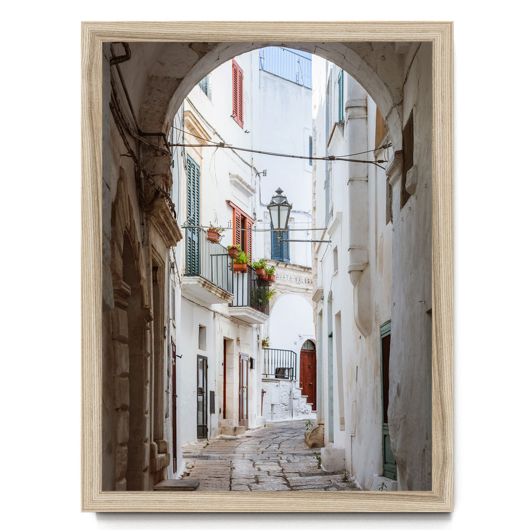 A narrow cobblestone street lined with white buildings and colorful shutters, leading to a balcony with potted plants.