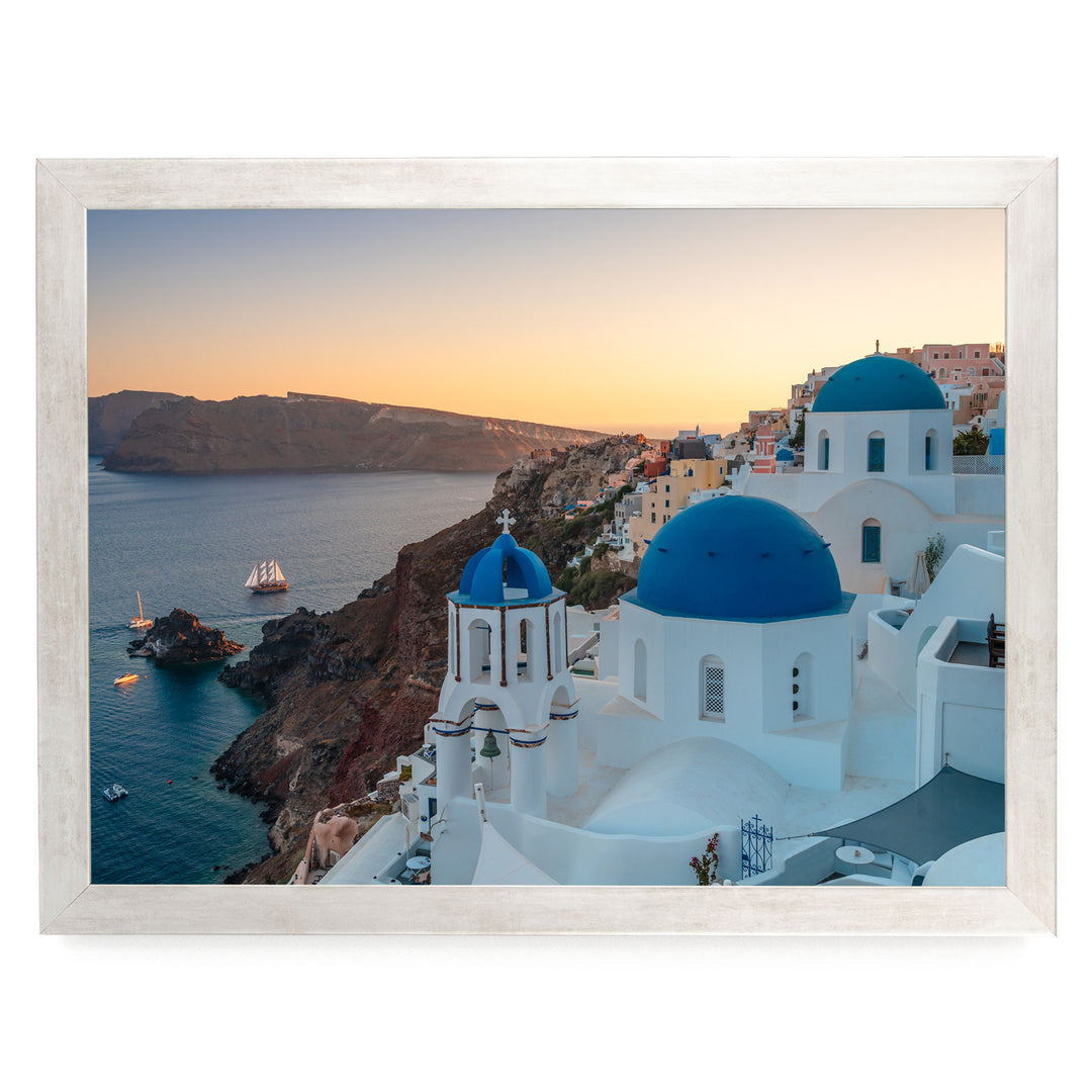 a picturesque coastal town with white buildings and blue domes, overlooking the ocean. A ship can be seen sailing in the distance, and the sky is painted in warm hues of orange, indicating the setting sun.