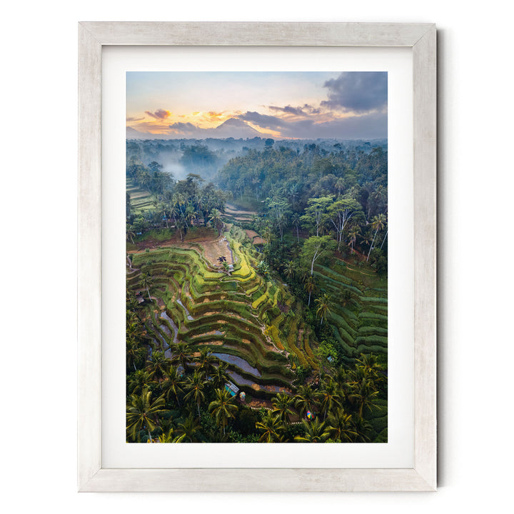 The image is a framed aerial view of a lush, terraced landscape with a mountain in the distance. The terraces are filled with vegetation, creating a verdant, tropical scene. The sky above is a beautiful gradient of orange and blue, suggesting either sunrise or sunset. The image is framed in a white wooden frame, which contrasts with the vibrant colors and natural elements of the scene.