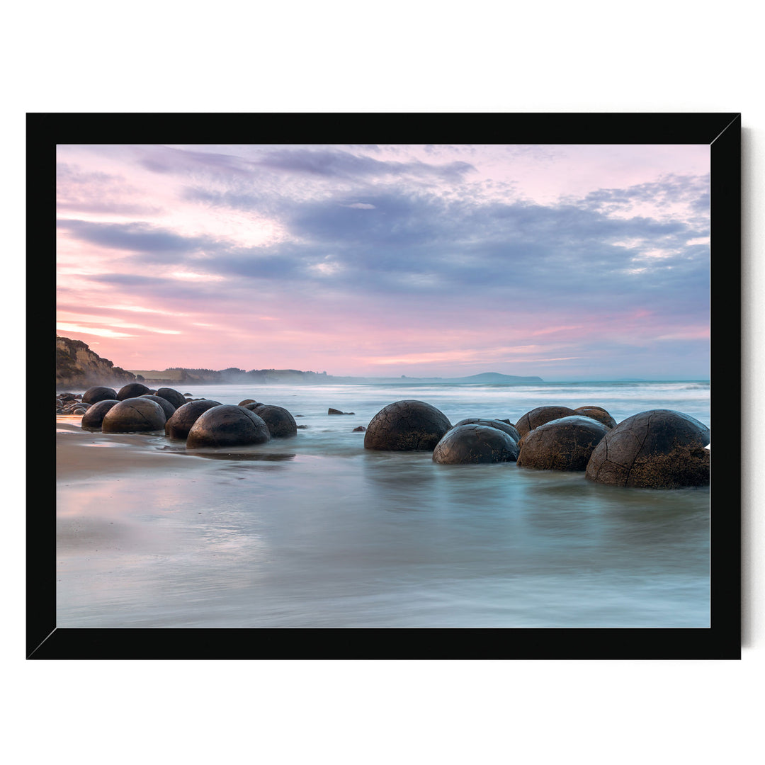 A serene beach scene with a collection of large, round boulders in the water, set against a backdrop of a colorful sunset sky.