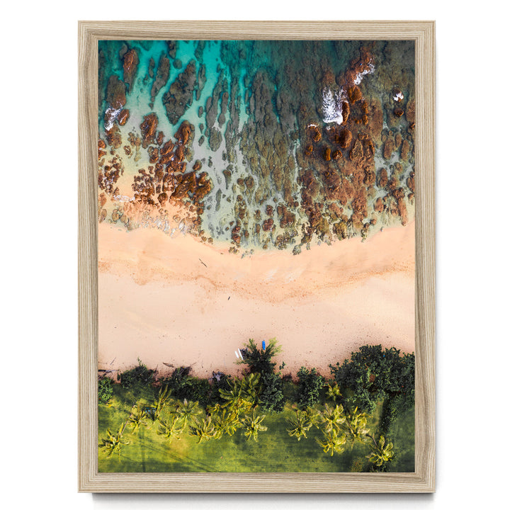 The image is a framed aerial view of a beach with a mix of sandy and rocky areas, surrounded by lush greenery and palm trees.