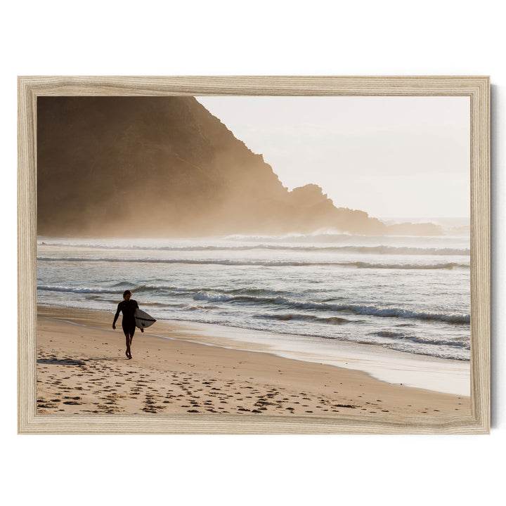 A person in a wetsuit carrying a surfboard walks along a sandy beach, with a rocky cliff and ocean in the background.