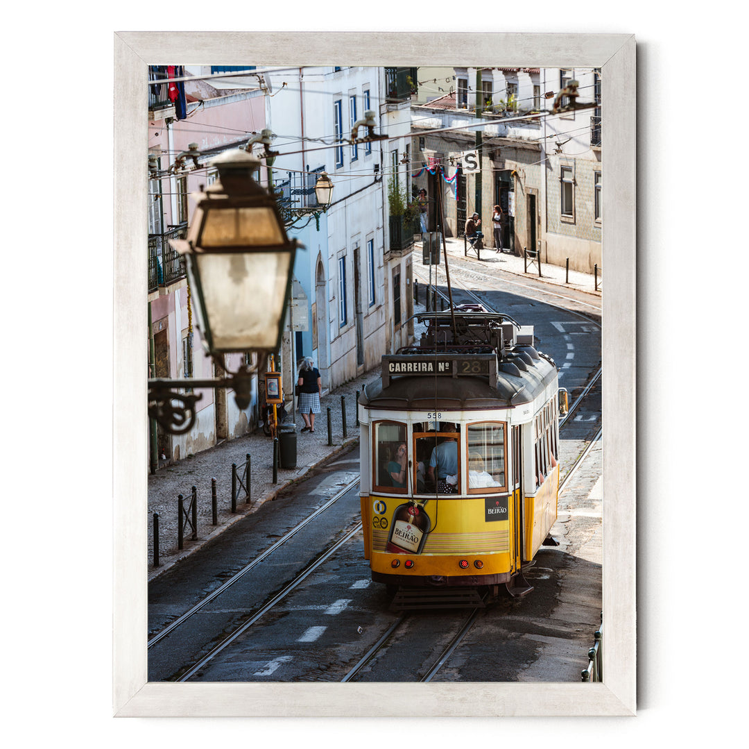 A yellow tram is traveling down a narrow street in a European city, with buildings and pedestrians visible in the background.