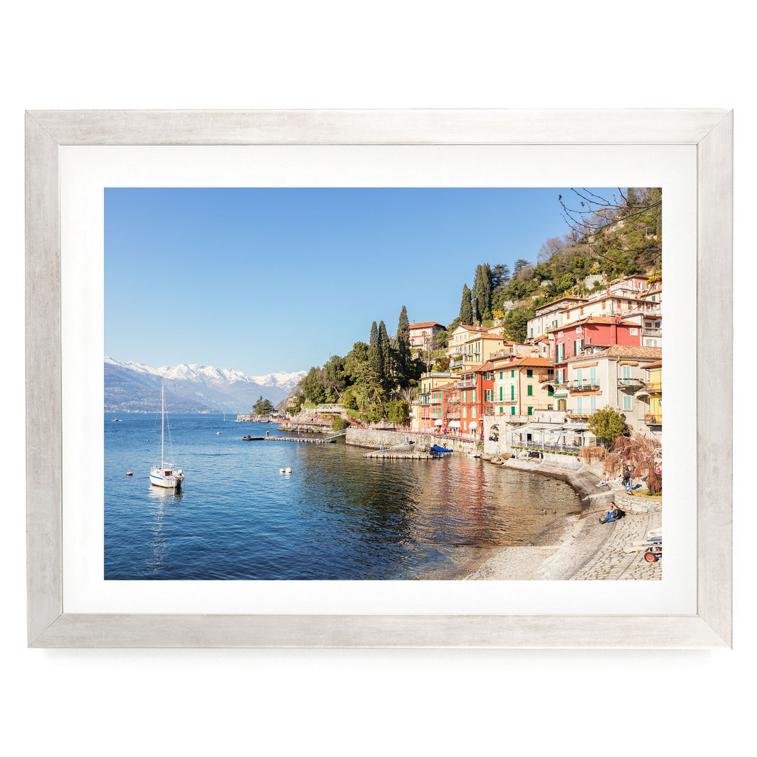 a picturesque coastal town with colorful buildings lining the shoreline, overlooking a calm lake. A sailboat is visible on the water, and the town is nestled among lush green hills.