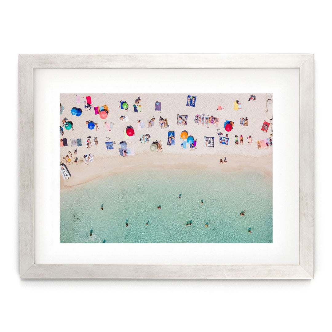 The image is a framed photograph depicting a crowded beach scene with numerous colorful umbrellas and people enjoying the water.