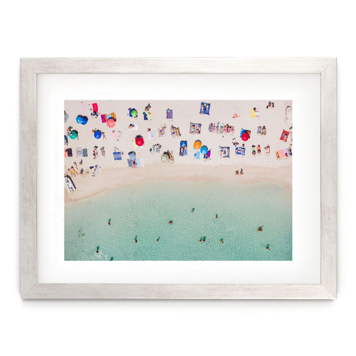 The image is a framed photograph depicting a crowded beach scene with numerous colorful umbrellas and people enjoying the water.