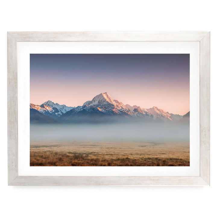 A breathtaking landscape featuring a majestic mountain range with snow-capped peaks, set against a vibrant sky at sunset. The foreground is a vast expanse of golden grass, creating a striking contrast with the natural scenery.