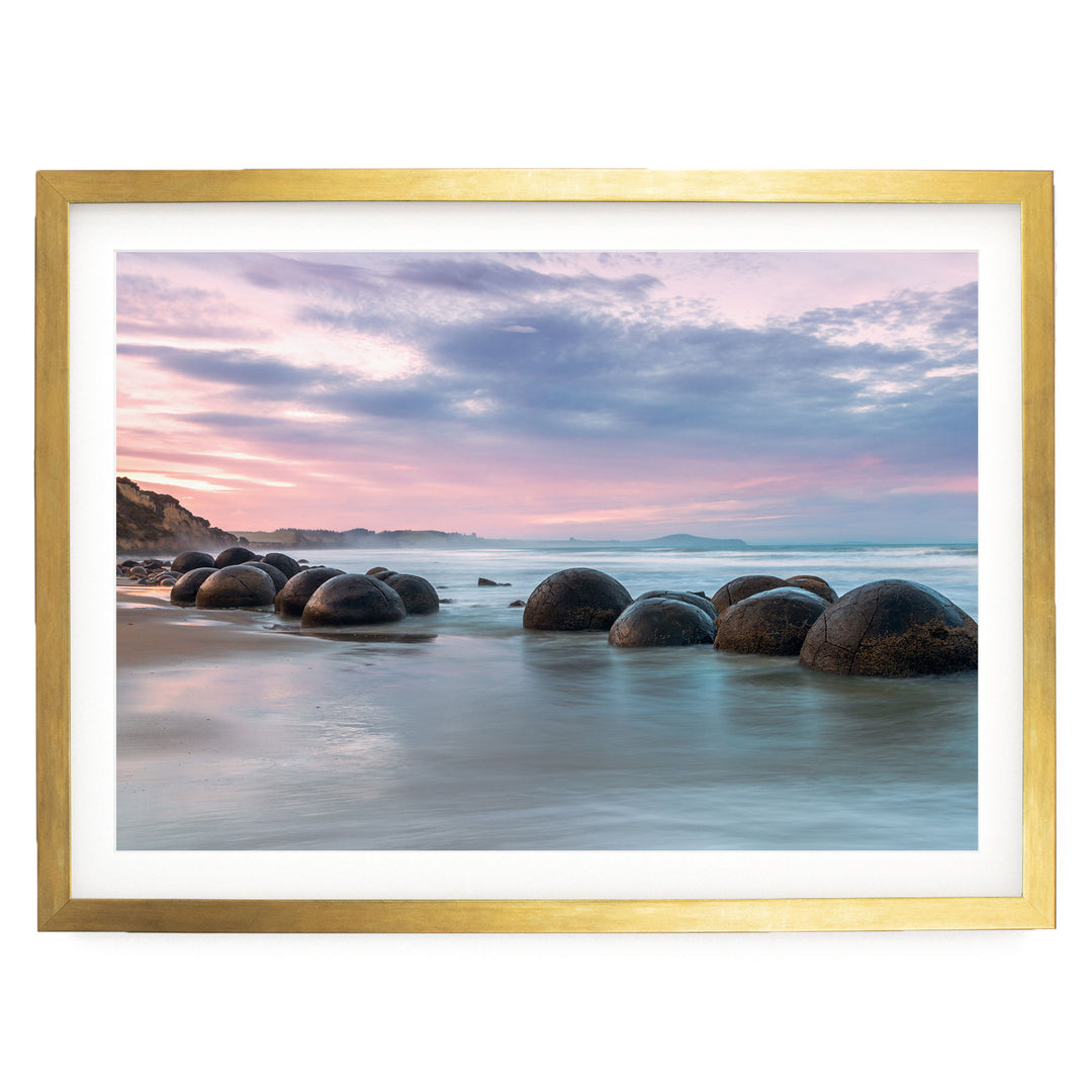 A serene beach scene with a collection of large, round boulders in the water, set against a picturesque sky with pink and purple hues.